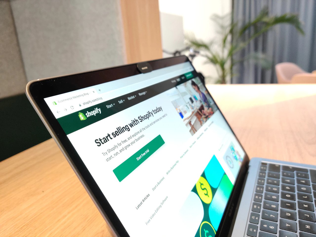 Ekranında Shopify sayfası açık olan bir laptop, laptobun arkasında bulanık arka planda bir bitki görünen, mobilyalı bir odada masanın üzerinde duruyor