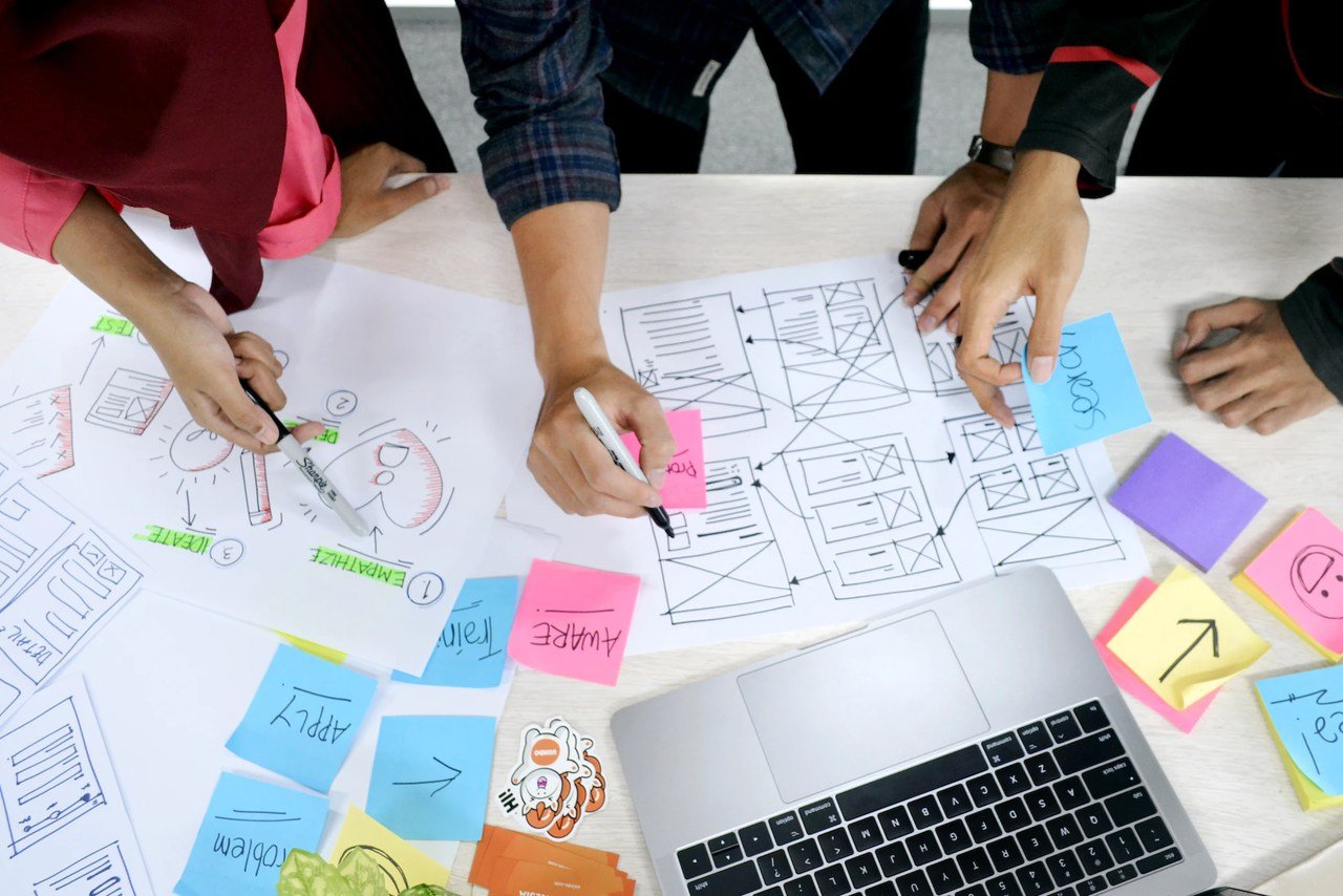 business strategy stock photo showing hands of three people some have pens showing each other somethings on a table with paper, stick notes and a laptop