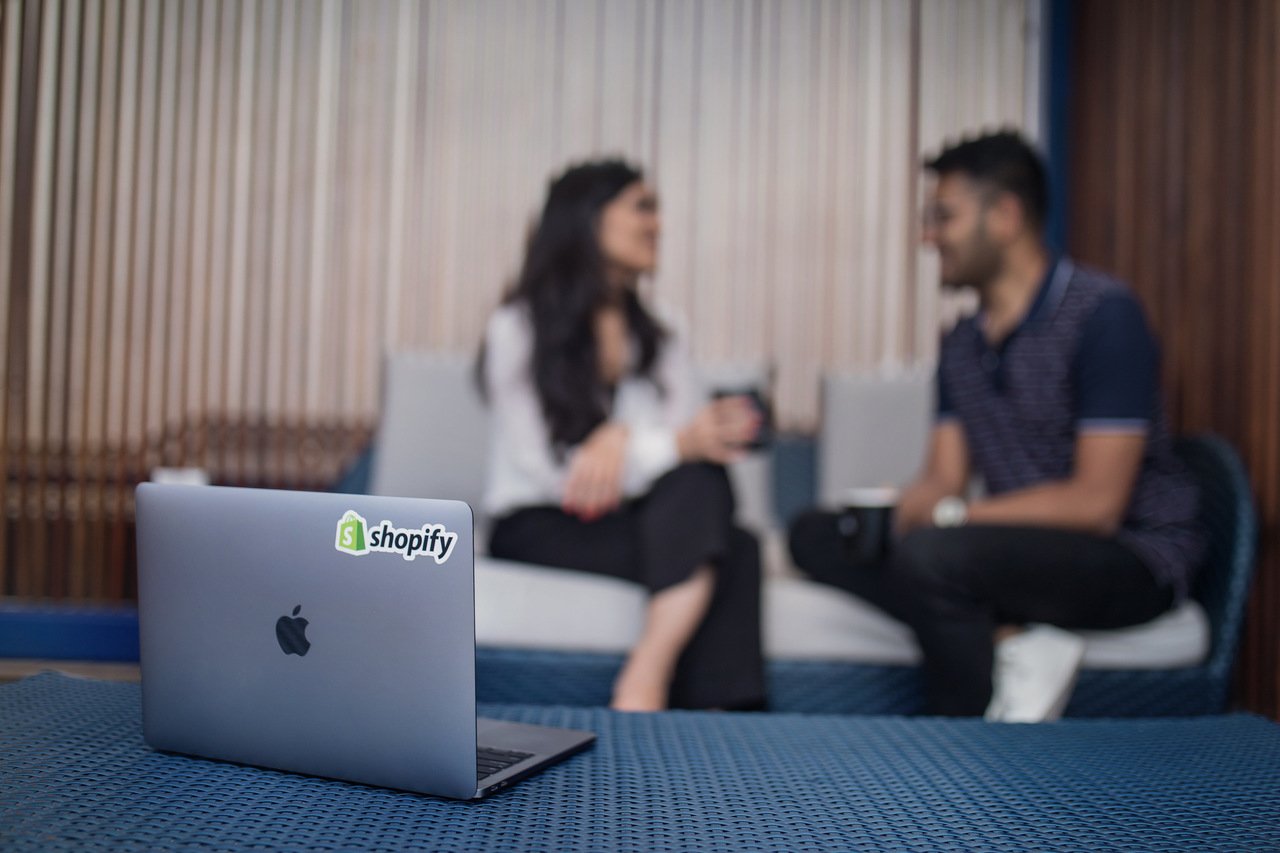 MacBook üzerinde Shopify çıkartması ve arka planda konuşan iki kişi