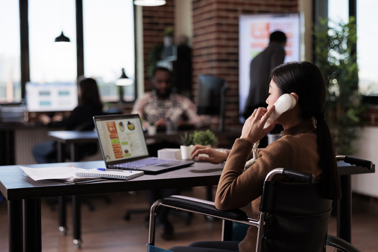 Bir kadın ofis masasında laptop ile telefon görüşmesi yapıyor; arka planda diğer insanlar ve bulanık ofis objeleri var