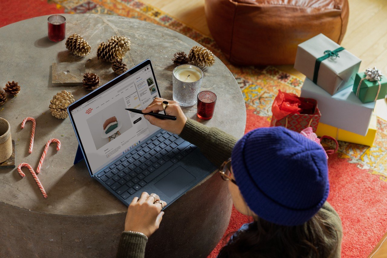 A person pointing at a laptop screen on a table; gift boxes are visible in the background