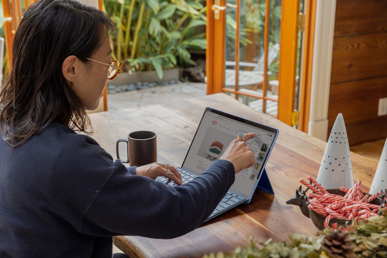 A person sitting at a desk points at the laptop screen with their finger; there is also a cup and some decorative items on the desk.