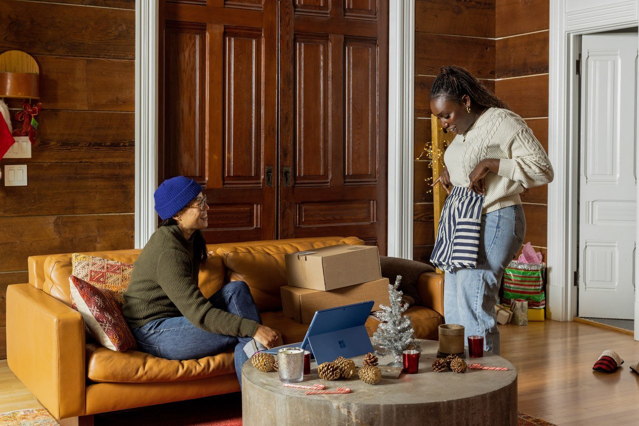 In a large room with wooden walls, a woman and a man; the woman is showing her clothes to the man, while the man is sitting on a sofa with shopping bags.