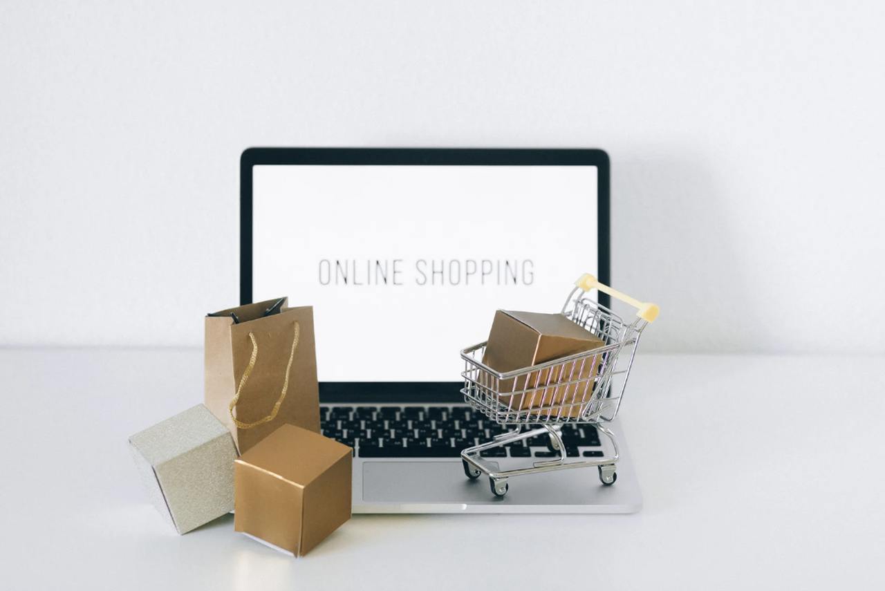 A MacBook displaying an online shopping page with small paper shopping bags and carts in front of it on a desk
