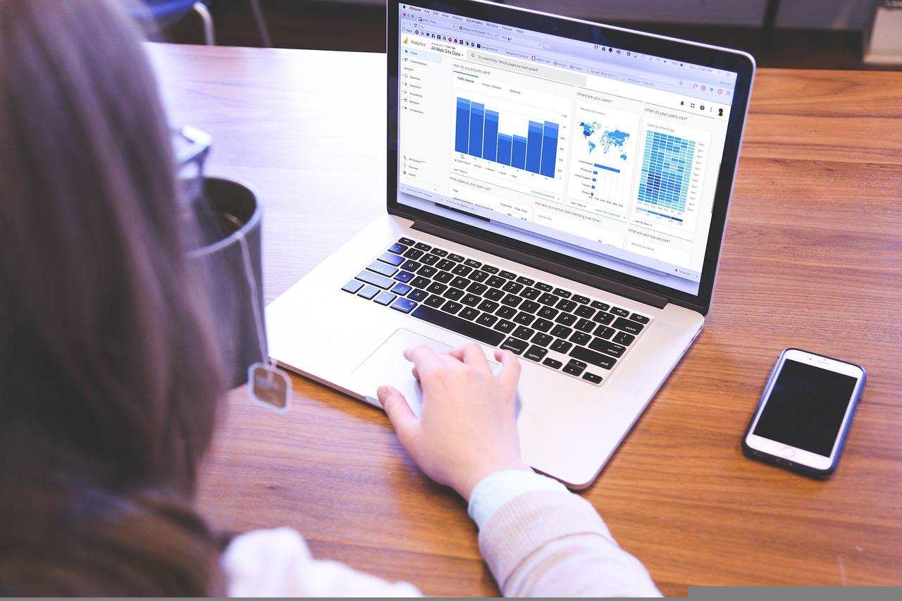 A woman working with Google Analytics, seen from behind, sitting at a desk with a phone on the right and a MacBook next to it