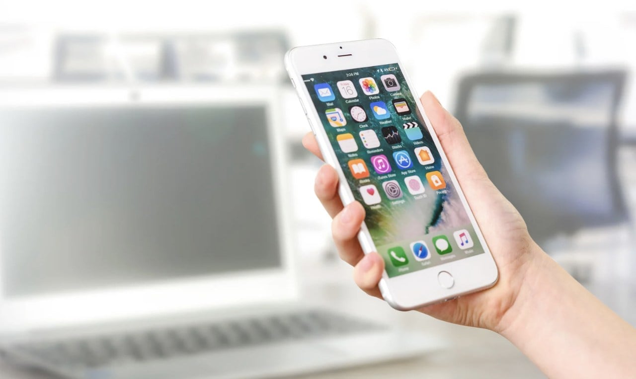 Hand holding smartphone showing installed apps with blurry background and laptop visible