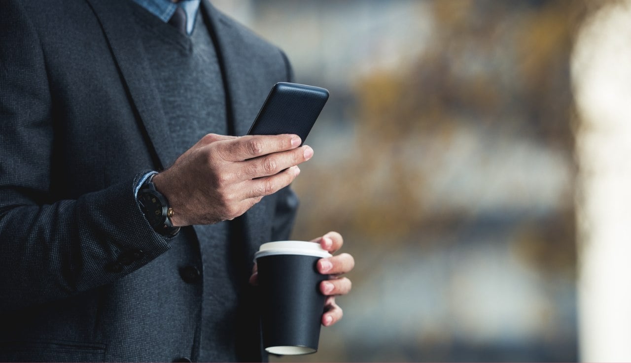 Siyah ceket giymiş bir iş adamı, bir elinde telefonunu kontrol ederken diğer elinde kahve bardağı tutuyor, bulanık bir arka planın önünde duruyor