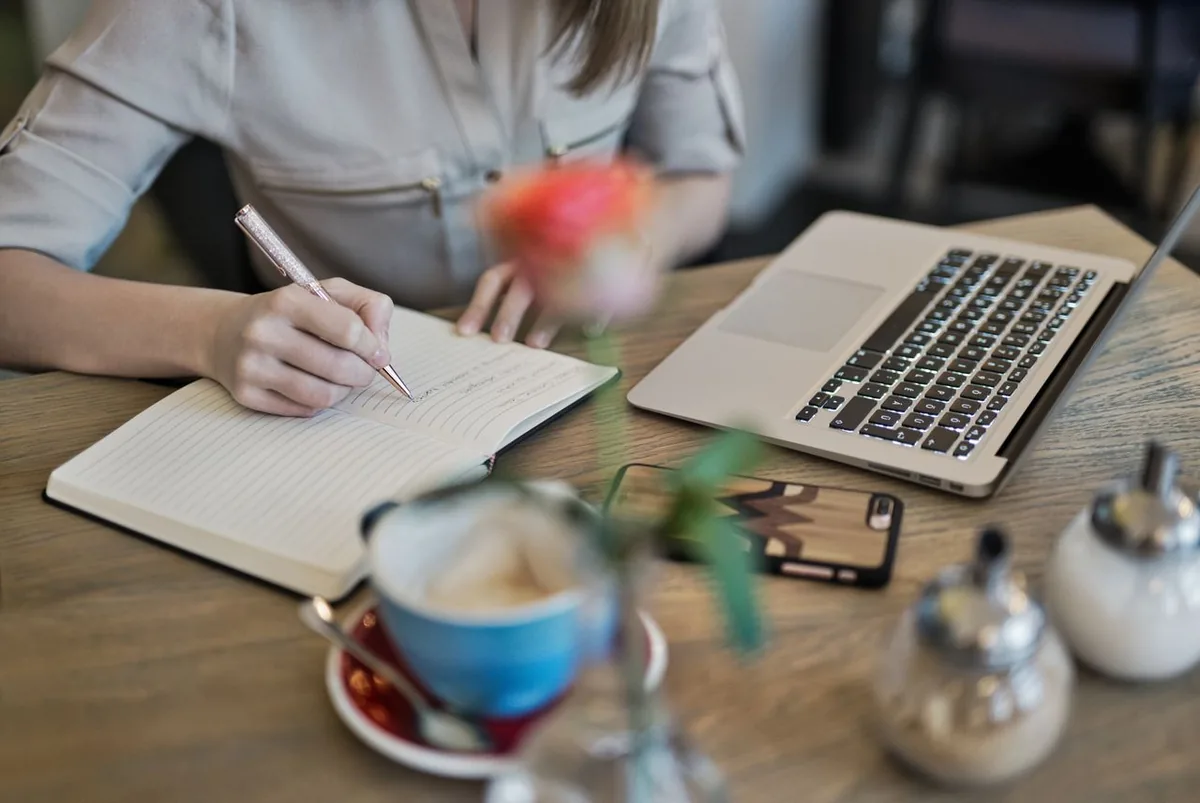 Person writing content in a notebook beside a laptop at a desk
